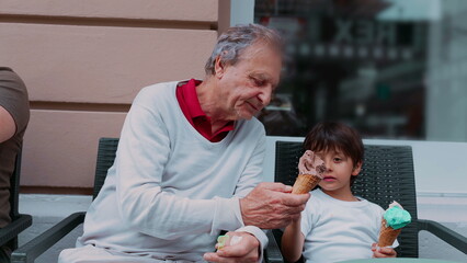 Young boy excitedly tasting his grandfather's ice cream while holding his own cone, capturing a joyful moment of sharing and connection between generations during a casual outdoor treat