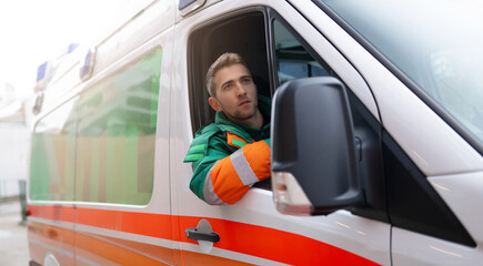 A male paramedic in a green and orange uniform is sitting in the driver's seat of an ambulance,...