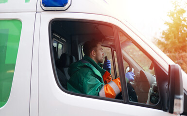 A male paramedic in a green and orange uniform is seated in an ambulance, holding a radio and communicating. He is prepared for emergency response, demonstrating readiness and professionalism.