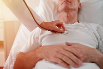Doctor listens to a patient's heartbeat with a stethoscope as the patient lies in bed. The patient’s hands rest on their chest, symbolizing trust, care, and medical attention in a healthcare setting