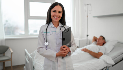 Smiling female doctor stands confidently in a hospital room with a clipboard and stethoscope. A...
