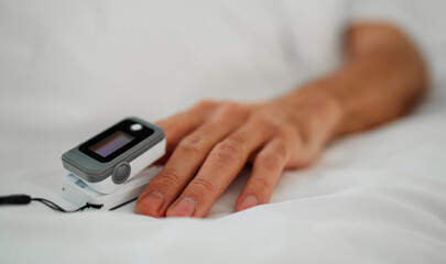 Close-up of a patient’s hand wearing a pulse oximeter, a medical device used to monitor oxygen levels and heart rate. The hand rests on a white hospital bed, highlighting health monitoring and care