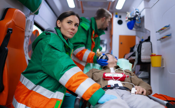 Paramedics in green and orange uniforms tend to male patient inside an ambulance. One monitors the patient while another provides oxygen. The scene highlights emergency medical response and teamwork - Powered by Adobe