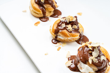 Profiteroles or eclairs poured with chocolate glaze with almond nut petals on a white plate and a light background. View from above. Space for text. Copyspace.