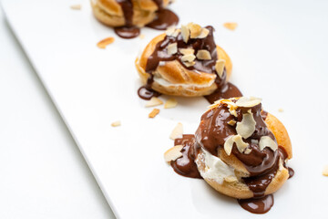 Profiteroles or eclairs poured with chocolate glaze with almond nut petals on a white plate and a light background. View from above. Space for text. Copyspace.