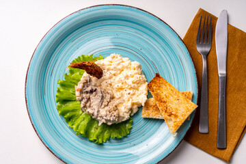 Scrambled eggs with mushrooms and crispy toast on a turquoise plate, decorated with lettuce, on a white table with a brown napkin and cutlery.