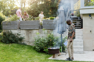 Man preparing grill for BBQ in backyard near blurred people on terrace