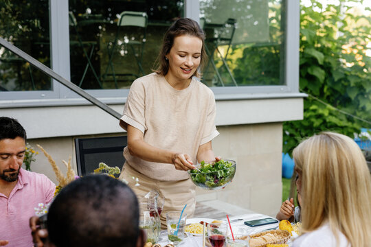 Woman putting salad from bowl to people having lunch at table