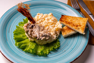 Scrambled eggs with mushrooms and crispy toast on a turquoise plate, decorated with lettuce, on a white table with a brown napkin and cutlery.