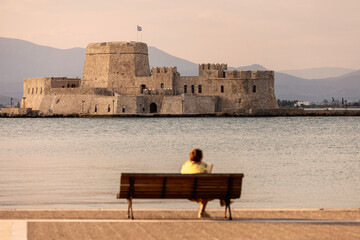 A solitary figure sits on a bench, gazing at the historic Bourtzi fortress in Nafplio, Greece, as the sun sets behind the serene waters, creating a tranquil atmosphere.