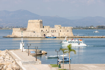 Bourtzi fortress stands majestically in the serene waters of Nafplio, surrounded by mountains. Local boats sail by, creating a picturesque coastal landscape that captures timeless beauty.
