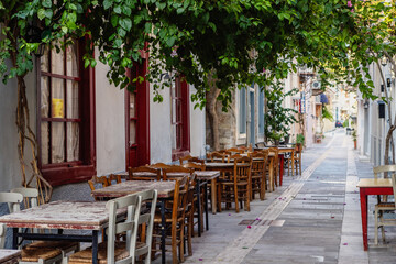 A serene street in Nafplio features rustic wooden tables lined under a canopy of lush vines, inviting guests to enjoy outdoor dining in a picturesque setting.