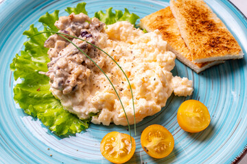 Scrambled eggs with mushrooms and crispy toast garnished with lettuce and yellow cherry tomatoes, on a turquoise plate, on a white table. Delicious and healthy breakfast. Close-up