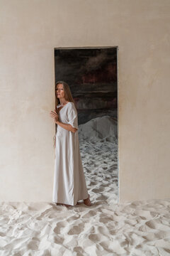 Woman in white dress standing in a sandy doorway with a serious expres