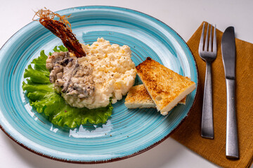 Scrambled eggs with mushrooms and crispy toast on a turquoise plate, decorated with lettuce, on a white table with a brown napkin and cutlery.