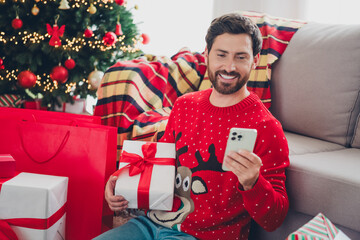 Portrait of nice young man use smart phone hold giftbox christmas holiday time flat indoors
