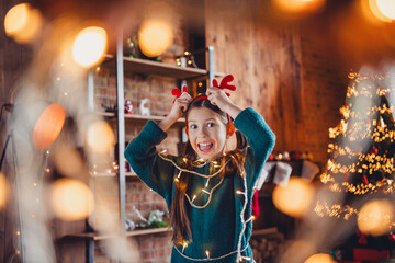 Photo of cute little girl excited wear deer horns have fun dressed green christmas outfit day light home party decoration interior room