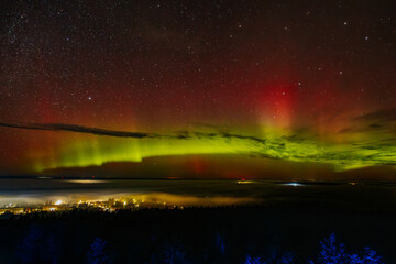 A vivid aurora borealis with striking red and green hues lights up the starry night sky above a town. The vibrant celestial display contrasts beautifully with the illuminated town below, Zilaiskalns.