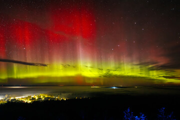 A vivid aurora borealis with striking red and green hues lights up the starry night sky above a town. The vibrant celestial display contrasts beautifully with the illuminated town below, Zilaiskalns.