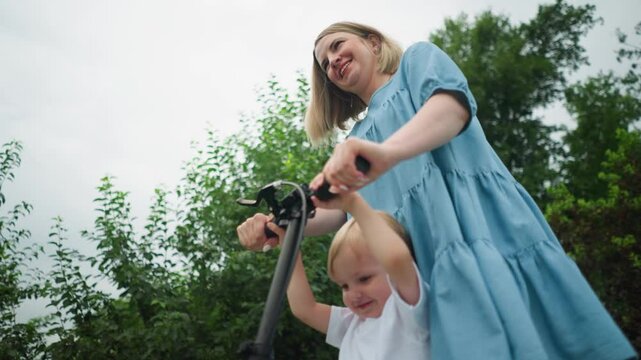 A mother and son ride a scooter together, both smiling warmly, close to a greenery background