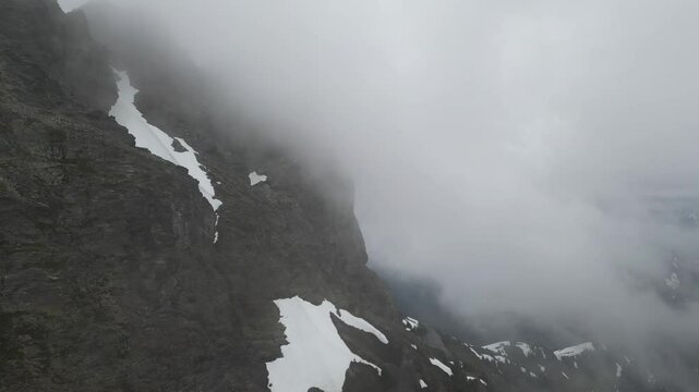 Aerial view of a steep, rocky mountain peak shrouded in thick fog, with patches of snow clinging to its slopes, evoking a sense of mystery and awe. British Columbia, Canada.