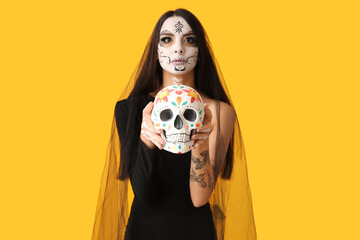 Young woman dressed as dead bride for Halloween party with painted human skull on yellow background