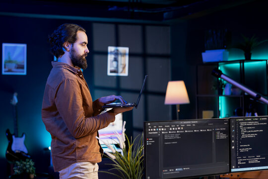 Developer doing software debugging using laptop, checking glitches in lines of code displayed on PC monitors. Man walking around home fixing coding issues using notebook device