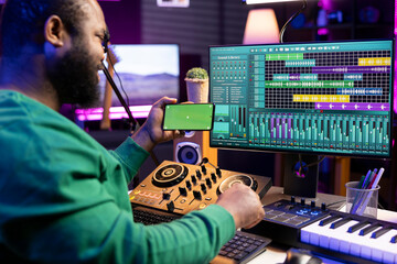 African american sound designer editing tracks with mixing console, looks at greenscreen on phone....