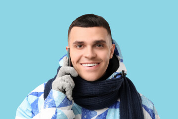 Young man in stylish puffer jacket and scarf talking by mobile phone on blue background