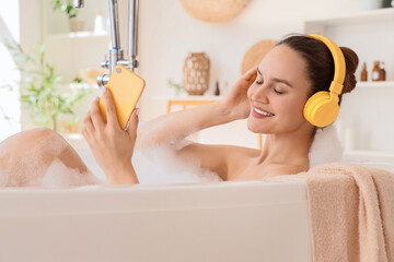 Naklejka premium Beautiful young happy woman in headphones holding mobile phone and taking bath with soap foam at home