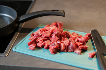 Close up of raw beef meat chopped in small cubes on green cutting board with a knife