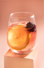 The image shows a clear glass filled with a refreshing drink, featuring a large slice of lemon and a few blackberries floating in the liquid. The glass is slightly covered with condensation droplets, 