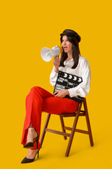 Young displeased female film director with movie clapper and megaphone screaming on chair against...