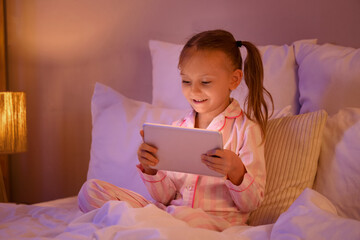 Cute happy little girl with tablet playing video games on bed in evening at home