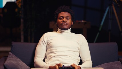 BIPOC player playing videogames on console late at night, struggling to stay awake on couch. Gamer using gamepad controller to try and finish game level before going to sleep, feeling sleepy