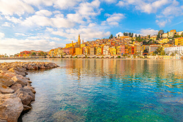 A rocky breakwater with the colorful old town, sandy beach, Mediterranean Sea and promenade behind, at Menton, France, along the Cote d'Azur French Riviera.