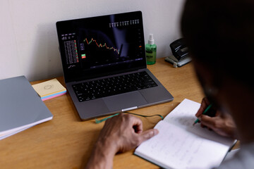 Partial man watching market graph on laptop and writing in notebook