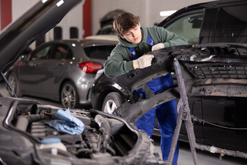 Young guy mechanic in uniform inspects car bumper in car service station