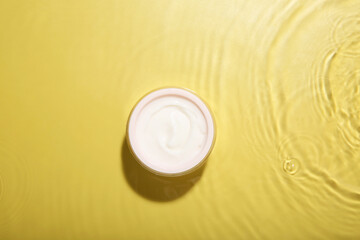 Jar with cream in water on yellow background, top view. Cosmetic product