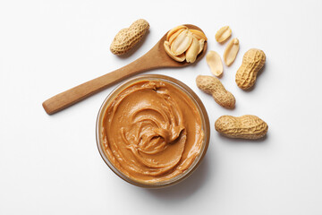 Tasty peanut butter in bowl and spoon with groundnuts on white table, flat lay