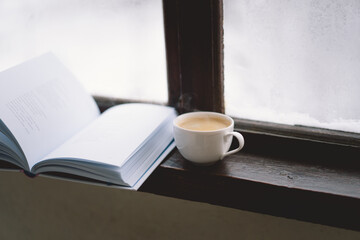 A warm cup of coffee rests beside an open book on a windowsill, while a lit candle flickers softly, creating a serene atmosphere against the snowy backdrop.