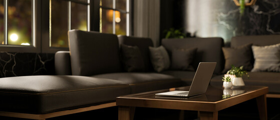 A laptop on a coffee table next to a large grey sofa in a modern black living room at night.