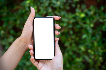 A close-up of a woman's hand holding a smartphone, set against a blurred background of green bushes.