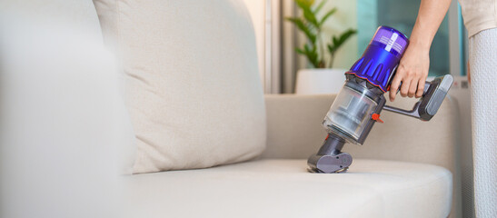 Woman cleaning Sofa with cordless Vacuum cleaner. Housewife using wireless Vacuum for big cleaning...