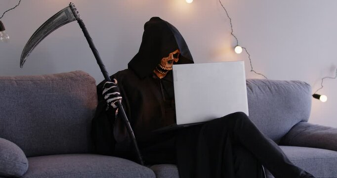 Grim reaper sitting on sofa in living room, using laptop computer, typing on computer keyboard, surfing internet. Sinister figure in hooded black cloak with scythe in his hand working online.