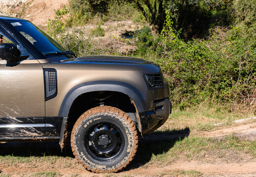 Close-up of the front right side view of a dark gold Land Rover New Defender with extreme mud tires driving on a dirt road