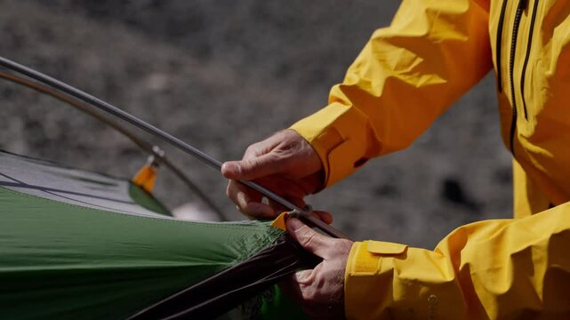 Closeup of a camper assembling a tent outdoors, sliding a tent pole through a fabric loop. The scene captures the details of setup in a sunny, rugged mountain environment