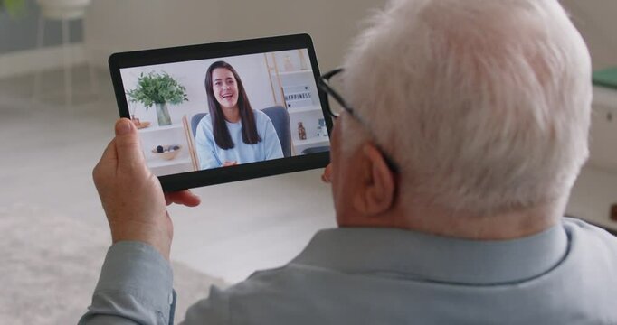 Portrait of an elderly senior man sitting on sofa in living room at home and having online video call with his adult daughter using tablet together. People and technology concept. 4k video.