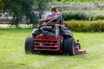 A person operating a lawn mower in a park, cutting the grass. The person is wearing a hat and headphones, focused on the task at hand