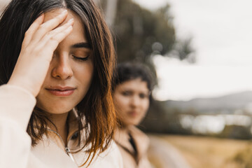 Close-up of a woman with her hand on her forehead, eyes closed, as another woman looks on in a natural outdoor setting